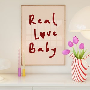 May include: A framed print with the text "Real Love Baby" in red lettering with a red heart in the word "Love". The print is on a pink background and is framed in a light wood frame. The print is sitting on a white dresser with two candles and a vase of pink tulips.