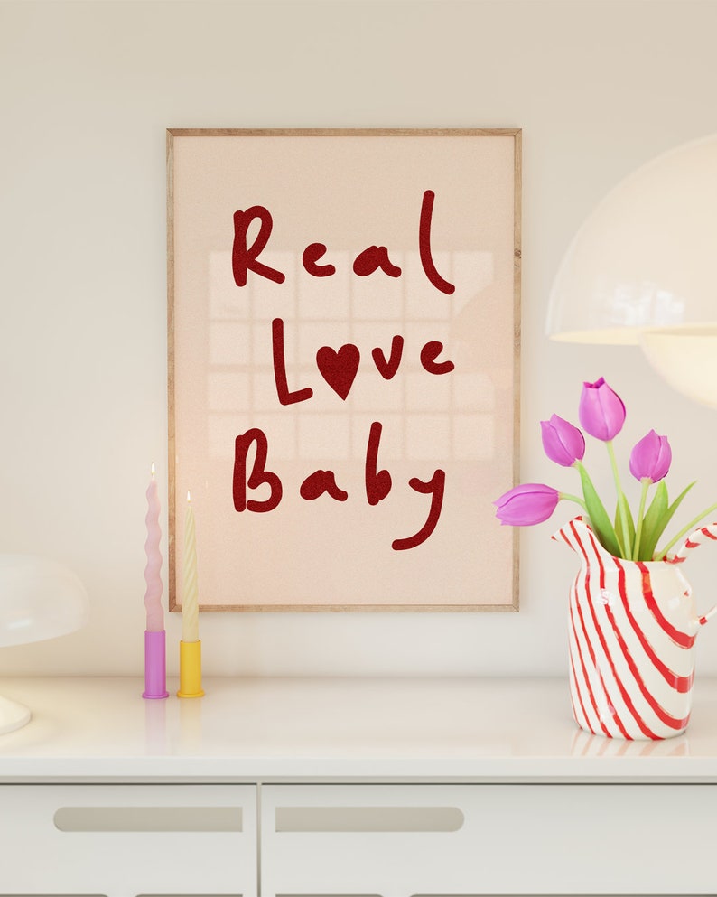 May include: A framed print with the text "Real Love Baby" in red lettering with a red heart in the word "Love". The print is on a pink background and is framed in a light wood frame. The print is sitting on a white dresser with two candles and a vase of pink tulips.