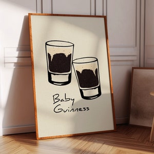 May include: A beige poster with a brown wood frame features a hand-drawn illustration of two small glasses of dark beer with a white head. The text "Baby Guinness" is written below the glasses.