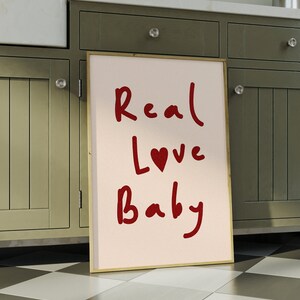 May include: A framed print with a pink background and red handwritten text that reads "Real Love Baby" with a red heart symbol in the word "Love".