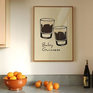 May include: A framed print with a white background and black line drawings of two shot glasses filled with a dark liquid. The text "Baby Guinness" is written below the glasses.