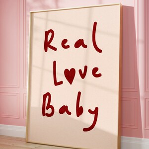 May include: A framed print with a pink background and gold frame. The print features the words "Real Love Baby" in red lettering with a red heart in the word "Love".