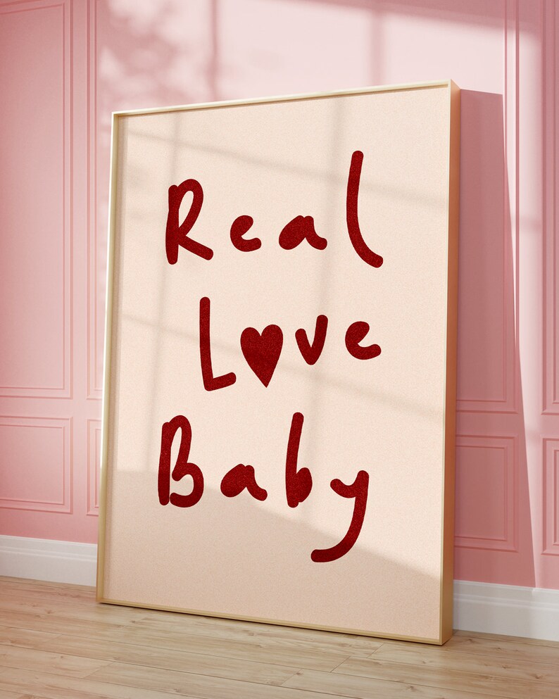 May include: A framed print with a pink background and gold frame. The print features the words "Real Love Baby" in red lettering with a red heart in the word "Love".
