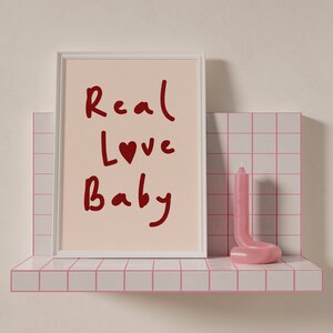 May include: A white picture frame with a pink background and the words "Real Love Baby" written in red. A pink candle is on a pink tiled shelf.