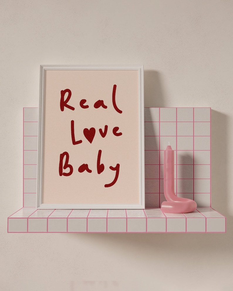 May include: A white picture frame with a pink background and the words "Real Love Baby" written in red. A pink candle is on a pink tiled shelf.