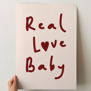 May include: A white poster with the words "Real Love Baby" written in red cursive. A small red heart is included between the words "Love" and "Baby".