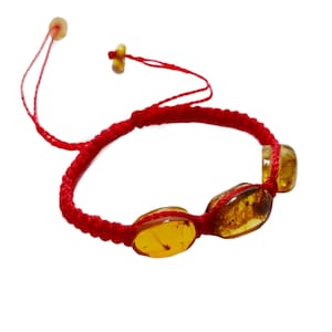 Puede incluir: Un brazalete de cuerda roja con tres cuentas de ámbar. Las cuentas son de forma ovalada y tienen una apariencia natural y translúcida.