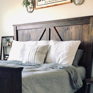 May include: A dark brown wooden headboard with a barn door style design. The headboard is on a bed with white pillows and a gray linen duvet.