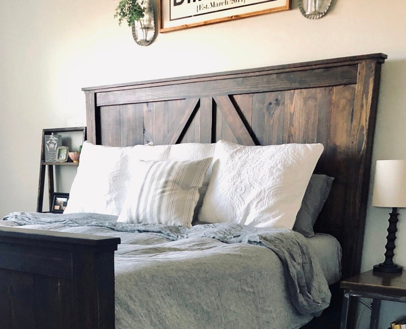 May include: A dark brown wooden headboard with a barn door style design. The headboard is on a bed with white pillows and a gray linen duvet.