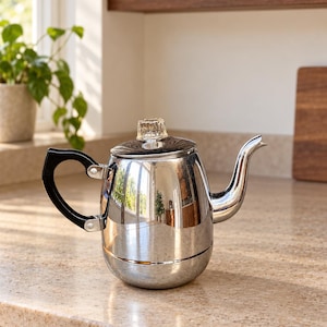 Könnte beinhalten: Eine polierte Chrom-Perkolator-Kaffeekanne mit gebogenem Ausguss, einem schwarzen, hitzebeständigen Griff mit silbernen Nieten und einem klaren Glasknopf auf dem Deckel. Die metallische Oberfläche reflektiert die Küchenumgebung.