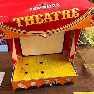 May include: A vintage View-Master Disney Theatre box set. The red box features the words "VIEW-MASTER" and "THEATRE" in yellow. The theater stage is yellow with black circles and a cartoon audience at the bottom. The set is designed to hold View-Master reels.
