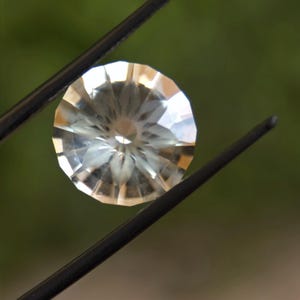 May include: A faceted, round gemstone held by black tweezers. The gem has a clear, pale yellow hue and a complex, symmetrical pattern of cuts that reflect light. The background is a soft, blurred green.