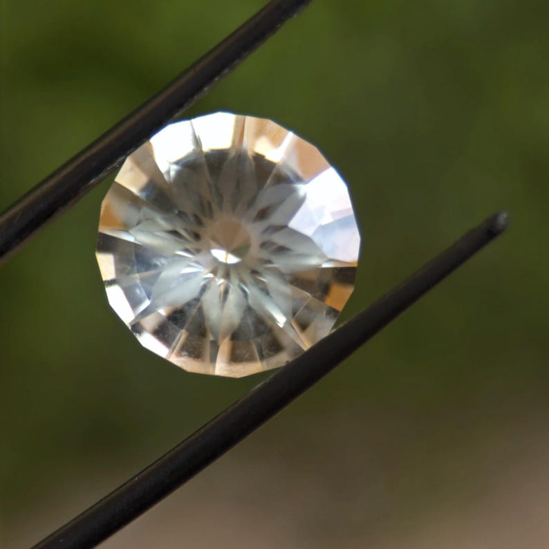 May include: A faceted, round gemstone held by black tweezers. The gem has a clear, pale yellow hue and a complex, symmetrical pattern of cuts that reflect light. The background is a soft, blurred green.