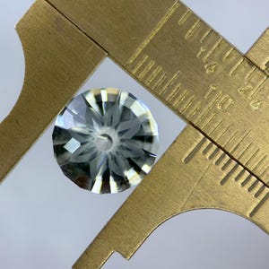 May include: A faceted, clear gemstone is measured with a brass caliper. The gemstone has a complex, starburst-like pattern of cuts. The caliper shows measurements in inches, with the gemstone appearing to be approximately 0.5 inches in diameter.