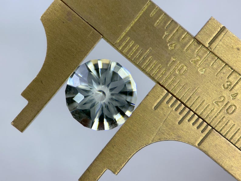 May include: A faceted, clear gemstone is measured with a brass caliper. The gemstone has a complex, starburst-like pattern of cuts. The caliper shows measurements in inches, with the gemstone appearing to be approximately 0.5 inches in diameter.