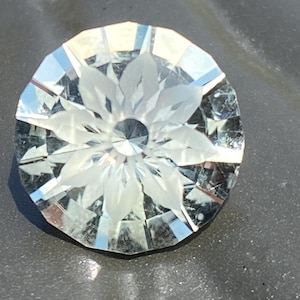 May include: A faceted, clear gemstone with a floral design carved into its center. The gem is round with many facets, reflecting light. The floral design is visible in the center of the gem.