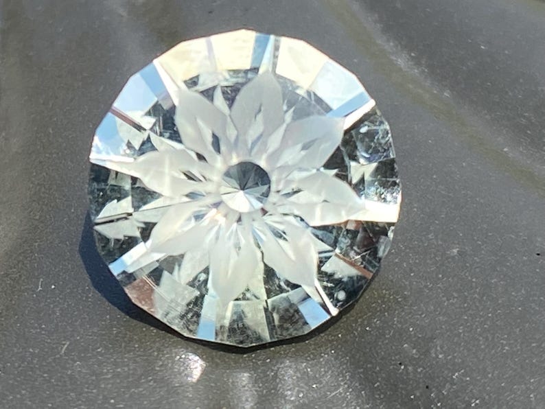 May include: A faceted, clear gemstone with a floral design carved into its center. The gem is round with many facets, reflecting light. The floral design is visible in the center of the gem.