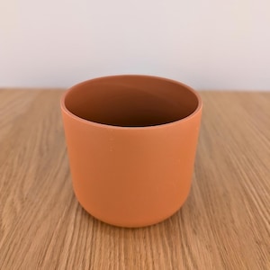 Peut inclure: Une tasse cylindrique de couleur terre cuite avec une finition mate lisse et une base arrondie. Le récipient présente une ouverture large sur le dessus et une teinte orange-brun solide et uniforme sur toute sa surface extérieure.