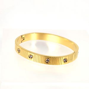 May include: A gold-colored bracelet with a hinged clasp. The bracelet features vertical grooved detailing and is adorned with several small, clear gemstones. The jewelry is displayed on a white surface.