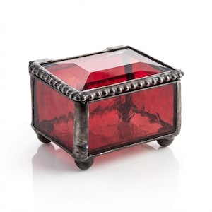 May include: A small, rectangular red glass jewelry box with a faceted lid. The box has a silver-colored metal frame and sits on four small, rounded feet. The edges are decorated with a beaded trim.