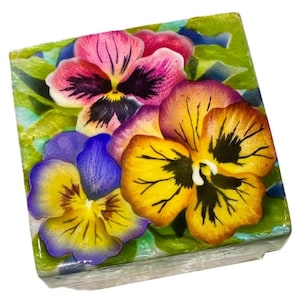May include: A square, decorative box featuring a vibrant floral design. The box is adorned with colorful pansies in shades of pink, yellow, and purple, set against a green and blue background. The flowers have detailed petals and dark centers.