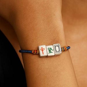 May include: A blue cord bracelet featuring three white rectangular charms with Chinese characters and symbols. The charms are connected by a blue cord with red and yellow accents. The bracelet is worn on a wrist.