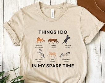 Könnte beinhalten: Ein beiges T-Shirt mit dem Text "THINGS I DO" und einer Liste von pferdebezogenen Aktivitäten, darunter "Zeit mit Pferden verbringen", "Pferde anschauen", "Pferde erforschen", "über Pferde sprechen", "Pferde füttern" und "von Pferden träumen". Der Text "IN MY SPARE TIME" befindet sich unter der Liste.