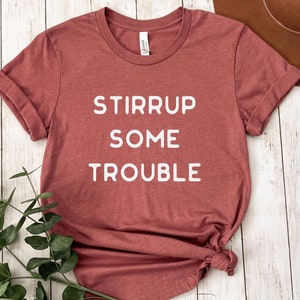 Puede incluir: Una camiseta color marrón rojizo con texto blanco que dice "Stirrup Some Trouble".