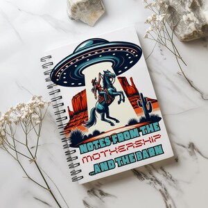 Puede incluir: Una libreta de espiral blanca con una ilustración colorida de una vaquera montando un caballo con un OVNI flotando arriba. El texto "Notes from the Mothership and the Barn" está impreso en la portada.