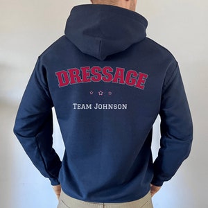 Puede incluir: Sudadera con capucha azul marino con el texto "Dressage" en rojo y "Team Johnson" en blanco. Hay tres estrellas rojas sobre el texto.
