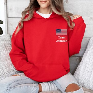 Puede incluir: Sudadera con capucha roja con una peque&ntilde;a bandera estadounidense y el texto "Team Johnson" en la parte delantera.