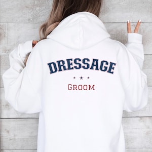 Puede incluir: Sudadera con capucha blanca con el texto "Dressage Groom" en letras azules y rojas en la parte posterior.