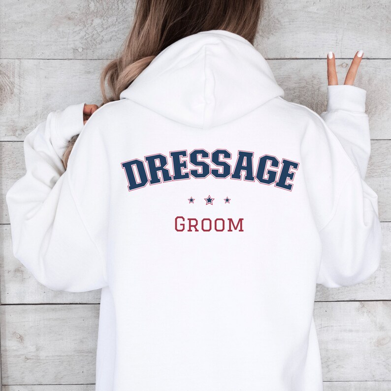 Puede incluir: Sudadera con capucha blanca con el texto "Dressage Groom" en letras azules y rojas en la parte posterior.