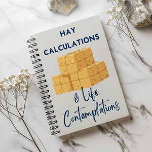 Puede incluir: Un cuaderno con espiral con las palabras "HAY CALCULATIONS & Life Contemplations" en azul marino. La portada presenta una ilustración de tres balas de heno amarillas y marrones. El cuaderno está sobre una superficie de mármol blanco.