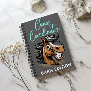 Puede incluir: Una libreta espiral gris con el texto "Chaos Coordinator: Barn Edition" en turquesa y blanco. Un caballo de dibujos animados con una sonrisa de dientes aparece en la portada.