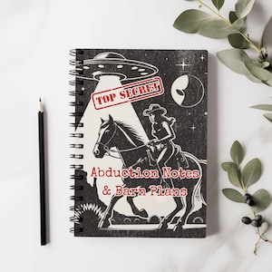 Puede incluir: Una libreta negra y blanca con encuadernación en espiral. La portada presenta a una vaquera montando a caballo con un OVNI en el fondo. El texto en la portada dice "Top Secret Abduction Notes & Barn Plans".