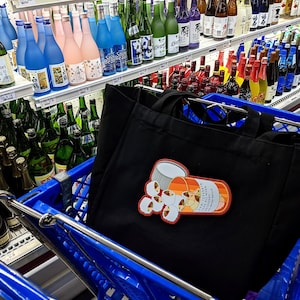 May include: A black tote bag with a colorful sticker of a pill bottle and pills inside a blue shopping cart, surrounded by shelves of various bottles of sake and other beverages.