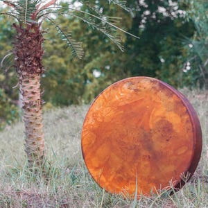 Pu&ograve; includere: Un grande tamburo rotondo e usurato, di colore marrone rossiccio, poggia su un pendio erboso. La superficie del tamburo mostra una trama screziata. Una palma si erge nelle vicinanze, con fogliame verde e un tronco strutturato.