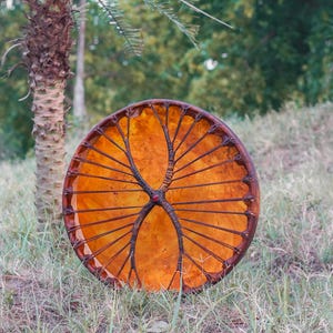 May include: A round, handcrafted drum with a warm orange-brown skin and a dark brown, tree-like design. The drum is set against a backdrop of green grass and a palm tree, suggesting an outdoor setting.