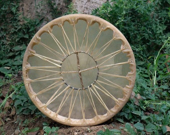 May include: A round, brown leather drum with a natural skin head. The drum is decorated with a cross pattern and has a series of leather laces around the edge.