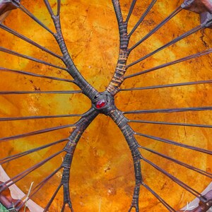 Pu&ograve; includere: Un tamburo rotondo fatto a mano con una pelle tesa giallo dorato. I raggi marrone scuro si irradiano da un punto centrale, adornato da una gemma rossa. Il telaio del tamburo &egrave; di un marrone pi&ugrave; scuro.