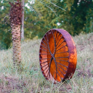 Pu&ograve; includere: Un grande tamburo rotondo con un telaio in legno marrone e una pelle tesa color arancione. Il tamburo &egrave; posizionato in posizione verticale in un campo erboso, con una palma e un fogliame verde sfocato sullo sfondo.