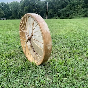 May include: A wooden frame drum with a natural leather drumhead and a branch woven into the frame. The drum is sitting on a grassy field.