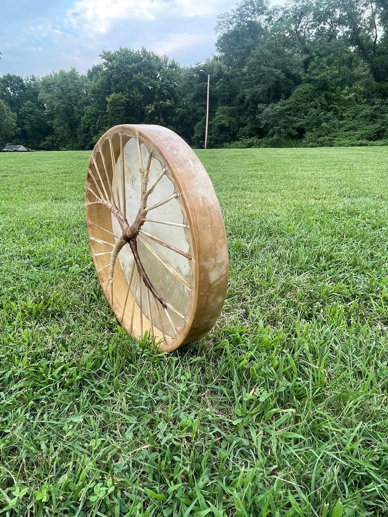 May include: A wooden frame drum with a natural leather drumhead and a branch woven into the frame. The drum is sitting on a grassy field.