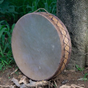 Puede incluir: Un tambor redondo de mano con un parche claro y un marco de madera. El marco está decorado con un patrón de diamantes repetido en una madera más oscura. Un asa de cuerda está unida en la parte superior. Mide unos 40 cm de diámetro.