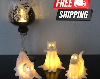 May include: A Halloween-themed display featuring three glowing ghost figurines and a glass candle holder. The candle holder has a black silhouette design of a haunted house, trees, and bats. The ghosts are white and lit from within.