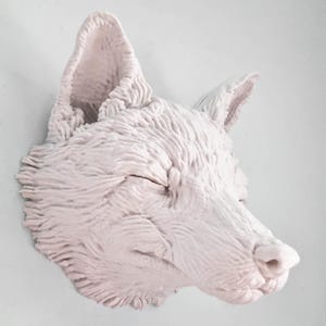 May include: A white ceramic fox head sculpture with closed eyes. The sculpture has a textured surface that resembles fur.