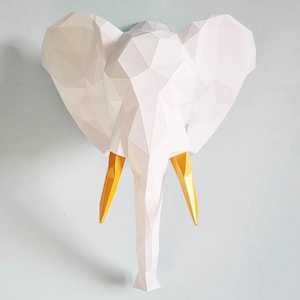 Könnte beinhalten: Weißer Origami-Elefantenkopf mit goldfarbenen Stoßzähnen. Der Elefant ist aus Papier gefertigt und hat ein geometrisches Design.
