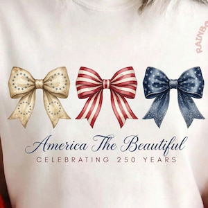 May include: Cream-colored t-shirt featuring three decorative bows in patriotic colors: tan with stars, red and white stripes, and navy with stars. The text "America The Beautiful Celebrating 250 Years" is printed below the bows.
