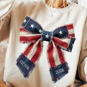 Op de afbeelding: Een crèmekleurige sweatshirt met een groot strikdesign in rood, wit en blauw, dat doet denken aan de Amerikaanse vlag. De strik bevat sterren, strepen en de tekst "EST. 1776" en "2026". De sweatshirt heeft een relaxte pasvorm.